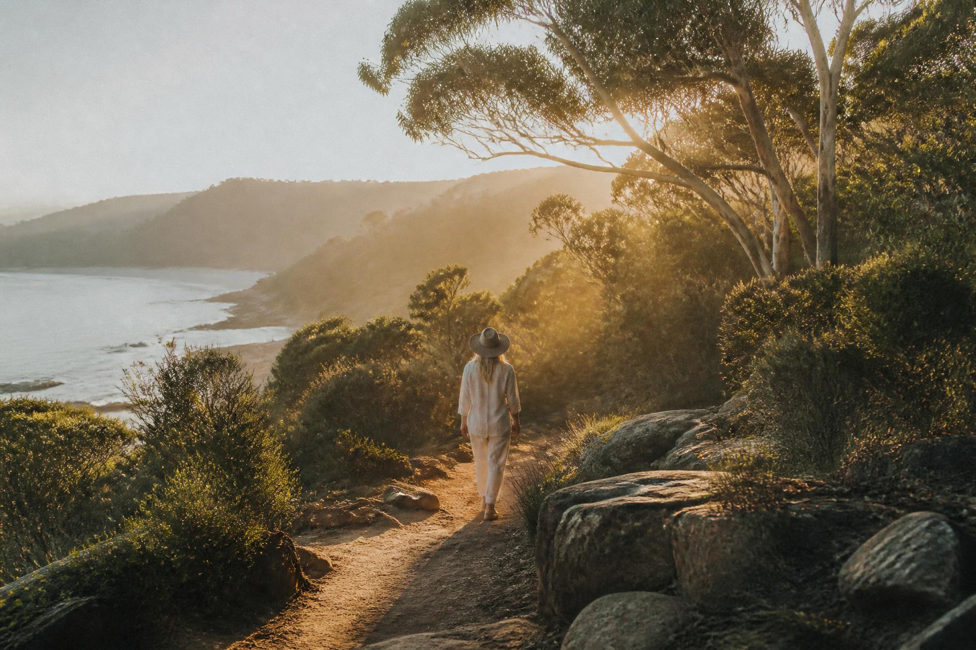 A woman walks a coastal path at sunrise beneath eucalyptus trees